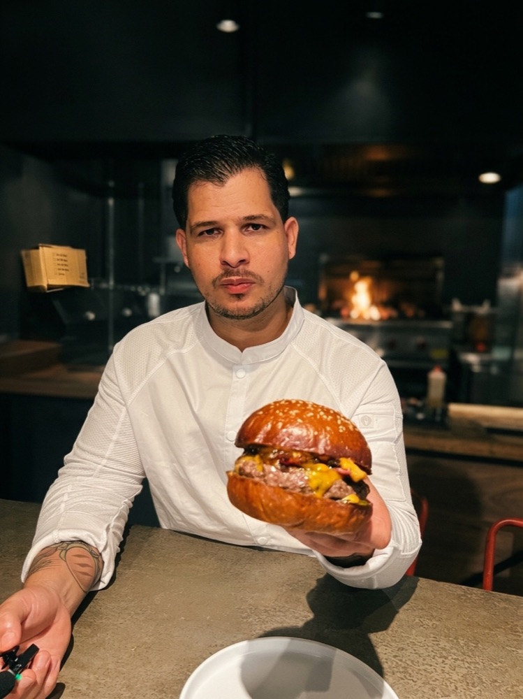 Chef Andrés Valerio holding a signature gourmet burger in his kitchen, warm low-key ambiance